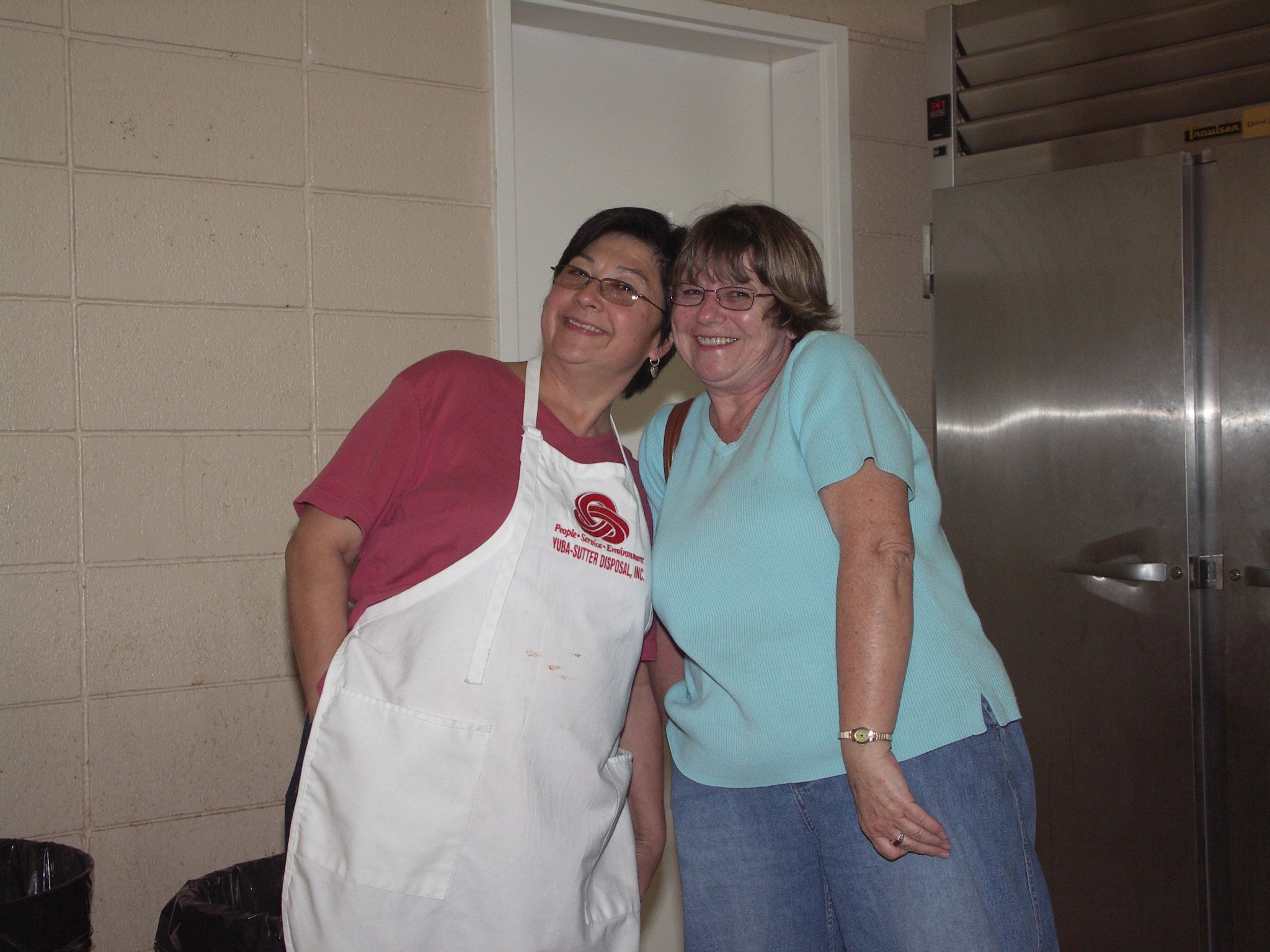 Two women smiling together near the church kitchen during a community dinner.